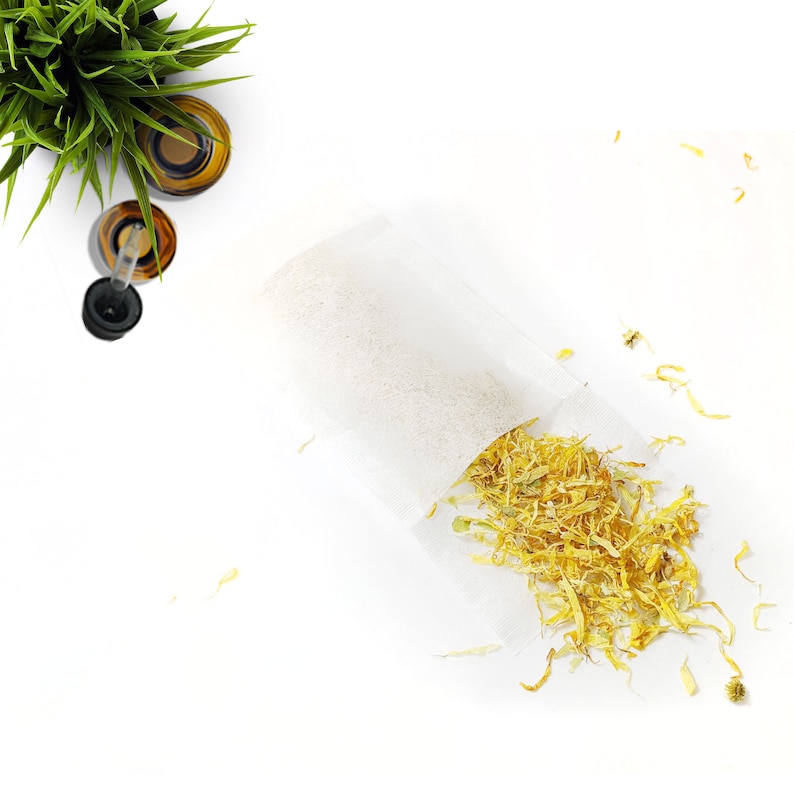 May include: Overhead shot of a white sachet filled with dried yellow flower petals, likely calendula, spilling onto a white surface. Two amber glass bottles with a dropper and a small green plant are in the upper left corner.