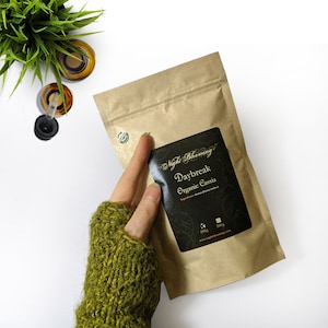 May include: A brown paper bag with a zip closure. The bag contains a product labelled "Daybreak Organic Cassia" with the brand name "Night Blooming". The label is black with gold lettering and features a small illustration of a flower.