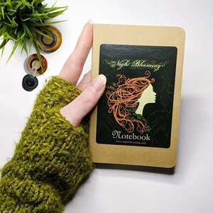 May include: A brown notebook with a sticker on the front. The sticker features a black background with a stylized illustration of a woman with orange hair and green leaves. The text on the sticker reads "Night Blooming" and "Notebook" with the website address "www.nightblooming.com".