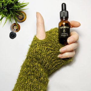 May include: A small amber glass bottle with a black dropper top and a black label that reads "Tenderhead Tranquility Oil" in gold lettering. The bottle is held in a hand wearing a green knitted mitten.