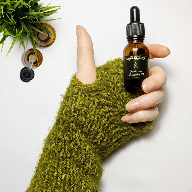 May include: A small amber glass bottle with a black dropper top and a black label that reads "Tenderhead Tranquility Oil" in gold lettering. The bottle is held in a hand wearing a green knitted mitten.