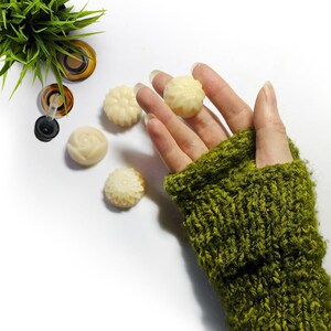 May include: Five small, white, round, decorative soap bars with floral designs. The soaps are on a white surface. A hand wearing a green knitted fingerless glove is holding one of the soaps.