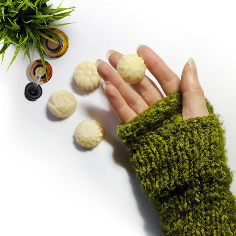 May include: Five small, white, round, decorative soap bars with floral designs. The soaps are on a white surface. A hand wearing a green knitted fingerless glove is holding one of the soaps.