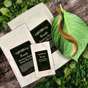 May include: Three brown paper packets with labels for Strawberry Blonde Herbal Hair Colour. The labels are black with gold lettering and feature a floral design. The packets are labelled with different weights: 100g, 100g, and 500g. A strand of light brown hair is on a green leaf.
