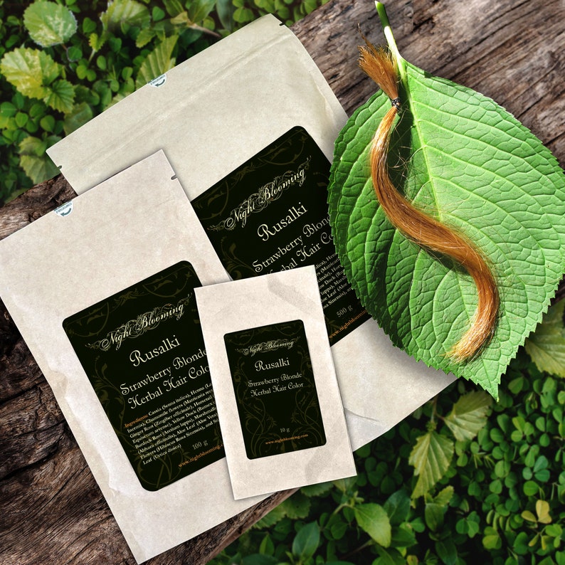 May include: Three brown paper packets with labels for Strawberry Blonde Herbal Hair Colour. The labels are black with gold lettering and feature a floral design. The packets are labelled with different weights: 100g, 100g, and 500g. A strand of light brown hair is on a green leaf.