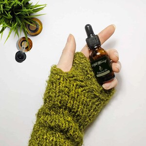 May include: A small amber glass bottle with a dropper top, labeled "Night Blooming Signature Oil". The bottle is held in a hand wearing a green knitted fingerless glove.