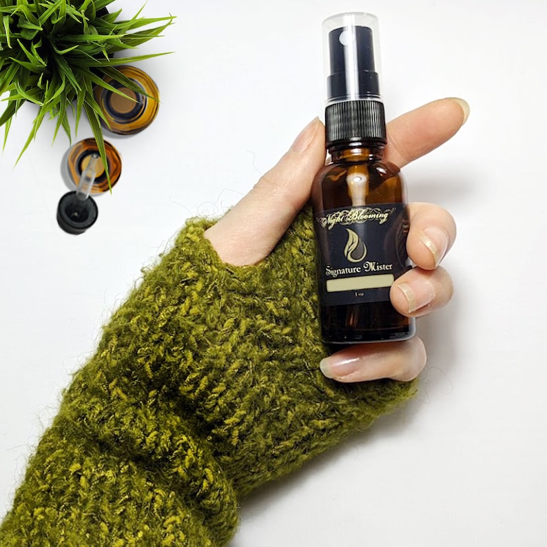 May include: A brown glass bottle with a black spray nozzle and a label that reads "Night Blooming Signature Mister". The bottle is being held by a hand wearing a green knitted mitten.