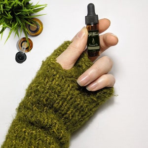 May include: A small brown glass bottle with a black dropper top and a label that says "Essential Oil". The bottle is being held by a hand wearing a green knitted fingerless glove.