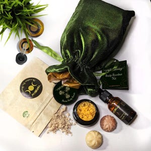 May include: A green velvet bag with a gold drawstring, a small brown paper bag with a label that says "Moonlit Shampoo", a small jar of yellow scrub with a label that says "Shrine Scalp Scrub", a small jar of brown powder, a small bottle of amber oil with a label that says "Scalp Oil", and two small round soaps. The label on the bag says "Scalp Soother Kit".