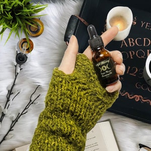 May include: A small amber glass bottle with a dropper, labeled "Triple Moon Anointing Oil" and "Witchy Alchemy". The bottle is held in a hand wearing a green knitted fingerless glove. The background includes a black tray with a Ouija board and a lit candle.