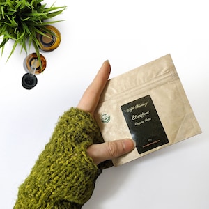 Puede incluir: Una bolsa de papel marrón con una etiqueta negra que dice "Light Blessings" y "Afterglow Organic Acids". La bolsa está siendo sostenida por una mano que lleva una manopla de punto verde.