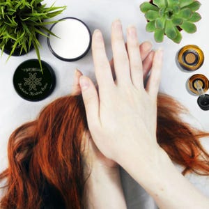 May include: A black jar with a gold label that reads "Featherless Healing Salve" with a stylized leaf design. The jar is on a white surface with a hand and red hair.