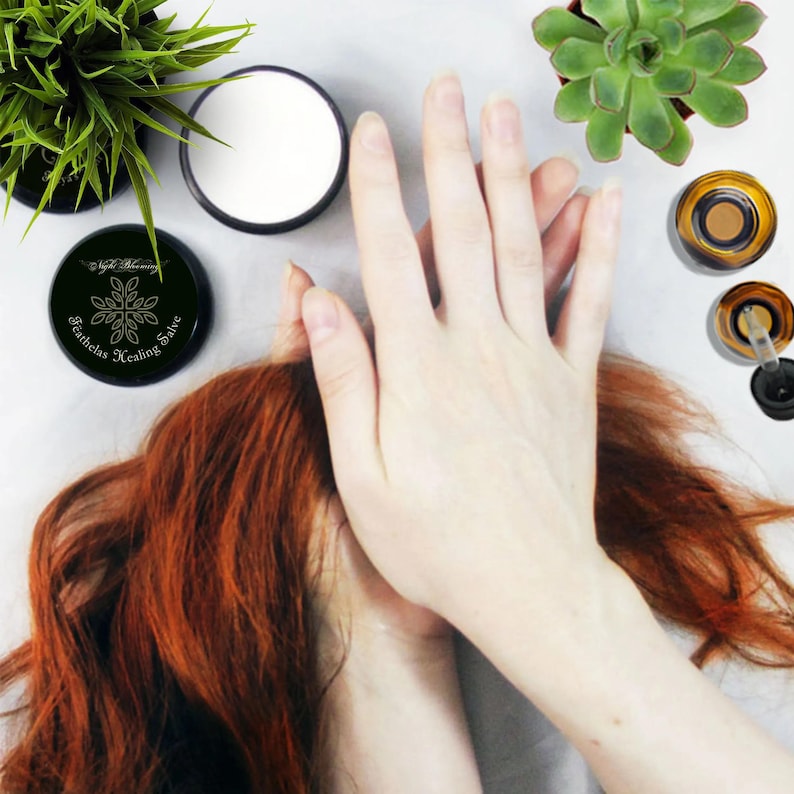 May include: A black jar with a gold label that reads "Featherless Healing Salve" with a stylized leaf design. The jar is on a white surface with a hand and red hair.