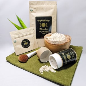 May include: A brown paper bag with the text "Night Blooming Moondust Dry Shampoo (Refill)" and a triple moon symbol. The bag is next to a wooden bowl filled with a light brown powder. A smaller brown paper bag with the same text and symbol is in the background. A white plastic container with a black label and the same text and symbol is open and spilling the powder onto a green cloth. A brown brush and a brown cardboard tube are also on the cloth.