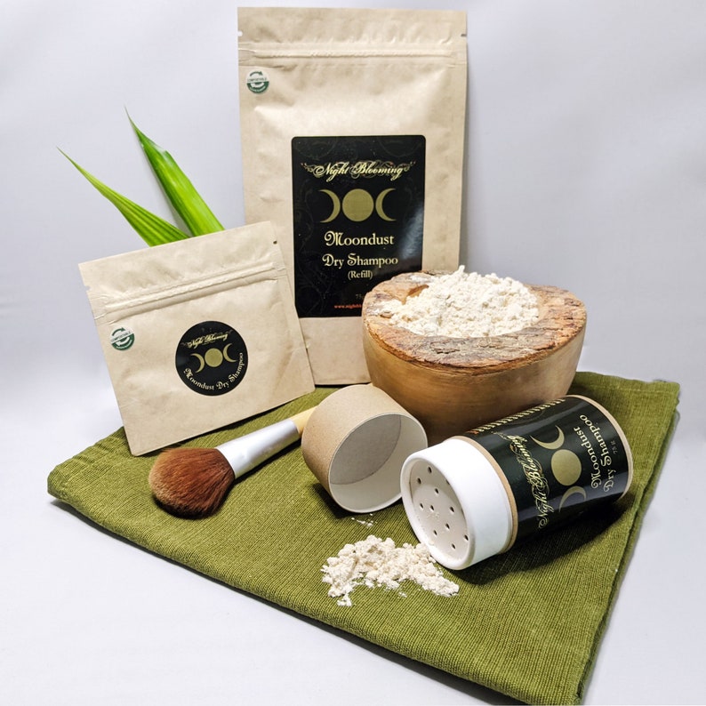May include: A brown paper bag with the text "Night Blooming Moondust Dry Shampoo (Refill)" and a triple moon symbol. The bag is next to a wooden bowl filled with a light brown powder. A smaller brown paper bag with the same text and symbol is in the background. A white plastic container with a black label and the same text and symbol is open and spilling the powder onto a green cloth. A brown brush and a brown cardboard tube are also on the cloth.