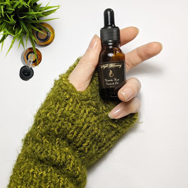Puede incluir: Una botella de vidrio marr&oacute;n con un gotero negro y una etiqueta dorada que dice "Kimochi Hair Growth Oil". La botella est&aacute; en una mano que lleva una manopla de punto verde.
