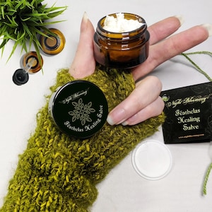 May include: A small amber glass jar with a white label that says "Night Blooming Feathalas Healing Salve" and a black label with the same text. The jar is open and contains a white salve. The jar is sitting on a green knitted glove.