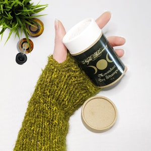 May include: A white container with a black label that says "Night Blooming Moondust Dry Shampoo 75g" with a crescent moon design. The container is being held by a hand wearing a green knitted fingerless glove.