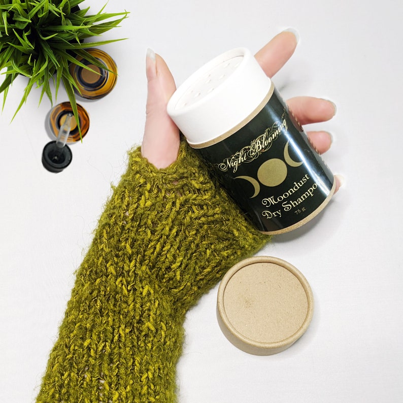 May include: A white container with a black label that says "Night Blooming Moondust Dry Shampoo 75g" with a crescent moon design. The container is being held by a hand wearing a green knitted fingerless glove.