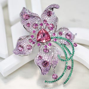 May include: A silver-toned flower brooch featuring a large triangular pink center stone, surrounded by numerous smaller pink and clear crystals. The petals are accented with pav&eacute;-set pink stones, while a curved stem is adorned with green crystals.