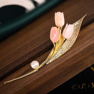 Puede incluir: Broche dorado con tres tulipanes de coral rosa. El broche tiene un diseño de hoja con cristales transparentes y una perla. Se encuentra sobre una superficie de madera oscura.
