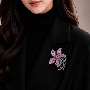 May include: A floral brooch featuring pink and purple petals with embedded crystals, accented by green curved stems. The brooch is pinned to the lapel of a black coat worn over a black turtleneck, creating a high-contrast accessory display.