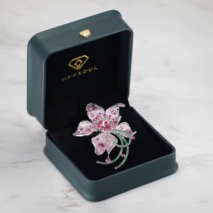 May include: A flower-shaped brooch featuring pink and clear crystals set in a silver-toned metal, displayed inside a dark green rectangular jewelry box with the gold-colored text CRYSOUL and a diamond logo printed on the interior lid lining.