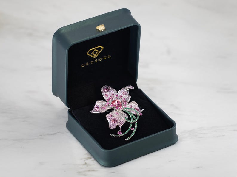 May include: A flower-shaped brooch featuring pink and clear crystals set in a silver-toned metal, displayed inside a dark green rectangular jewelry box with the gold-colored text CRYSOUL and a diamond logo printed on the interior lid lining.