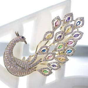 May include: A gold-coloured peacock brooch adorned with sparkling crystals and colourful gemstones. The brooch features a detailed peacock design with a curved neck, a fan of feathers, and various coloured gems.