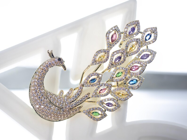 May include: A gold-coloured peacock brooch adorned with sparkling crystals and colourful gemstones. The brooch features a detailed peacock design with a curved neck, a fan of feathers, and various coloured gems.