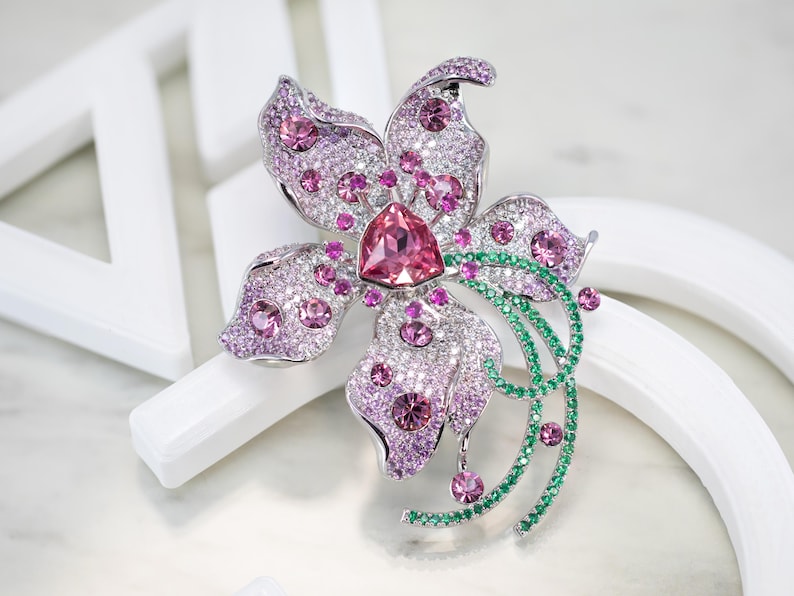 May include: A silver-toned flower brooch featuring a large triangular pink center stone, surrounded by numerous smaller pink and clear crystals. The petals are accented with pav&eacute;-set pink stones, while a curved stem is adorned with green crystals.