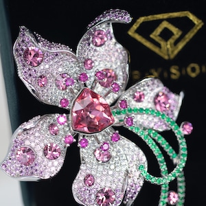 May include: A silver-toned flower brooch featuring a large central triangular pink crystal, surrounded by smaller round pink crystals and pav&eacute;-set clear stones. The design includes curved stems accented with small green crystals and a gold logo.