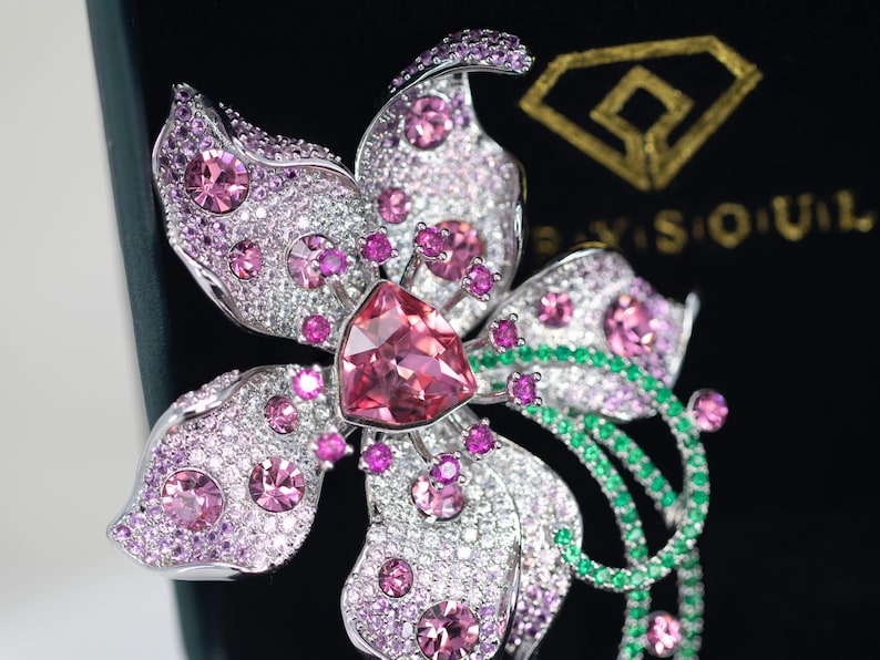 May include: A silver-toned flower brooch featuring a large central triangular pink crystal, surrounded by smaller round pink crystals and pav&eacute;-set clear stones. The design includes curved stems accented with small green crystals and a gold logo.