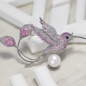 May include: A silver hummingbird brooch with pink and white crystals and a pearl accent. The hummingbird is perched on a branch with two leaves.