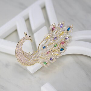 May include: A gold peacock brooch, its body and head covered in clear crystals. The tail feathers feature colourful gemstones in blue, yellow, pink, and purple. The brooch is displayed against a white backdrop.
