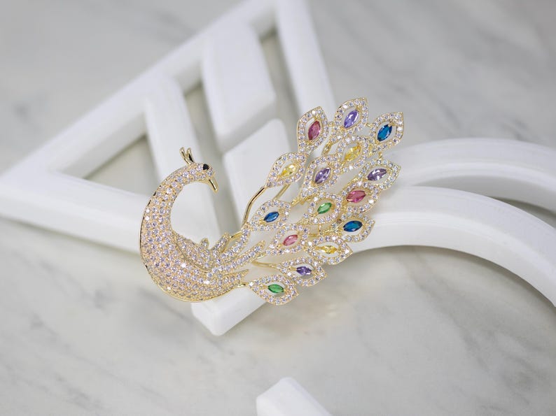 May include: A gold peacock brooch, its body and head covered in clear crystals. The tail feathers feature colourful gemstones in blue, yellow, pink, and purple. The brooch is displayed against a white backdrop.