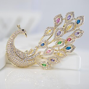 May include: A gold peacock brooch, adorned with clear and coloured gemstones. The brooch has a detailed design, with a curved neck, a body covered in small stones, and a fanned tail with various coloured gems.
