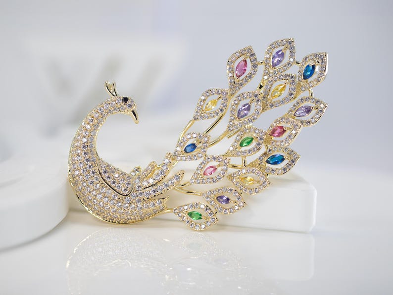 May include: A gold peacock brooch, adorned with clear and coloured gemstones. The brooch has a detailed design, with a curved neck, a body covered in small stones, and a fanned tail with various coloured gems.