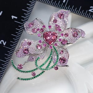 May include: A silver-toned flower brooch featuring a large triangular pink center stone, surrounded by numerous small pink and clear crystals. The brooch includes curved stems accented with green crystals and measures approximately 2 inches wide.