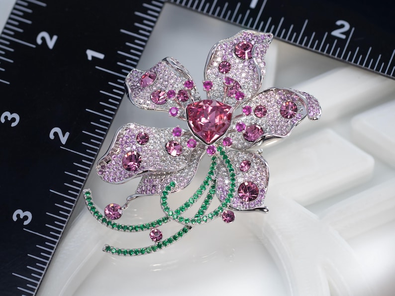 May include: A silver-toned flower brooch featuring a large triangular pink center stone, surrounded by numerous small pink and clear crystals. The brooch includes curved stems accented with green crystals and measures approximately 2 inches wide.