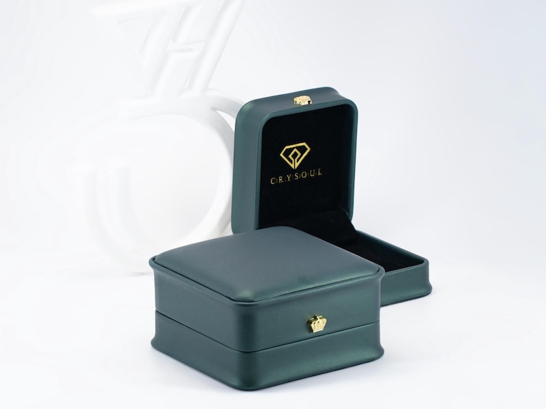 May include: Two dark teal jewellery boxes, one open and one closed, with gold-coloured accents. The open box reveals a black interior with gold text and a diamond-shaped logo. The closed box has a gold crown-shaped clasp.