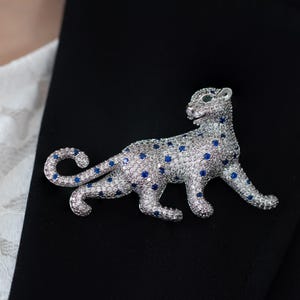 May include: A silver-toned leopard brooch with a sparkling design. The brooch features a detailed leopard shape, covered in small, clear crystals with blue accents. The leopard is posed in a walking position, with green eyes.