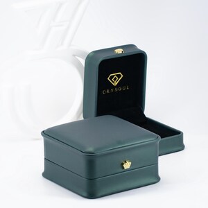 May include: A dark green square jewelry box with a smooth finish and a gold crown-shaped clasp on the front. The interior is lined with black velvet and features a gold diamond-shaped logo with the text CRYSOUL printed on the inside lid.