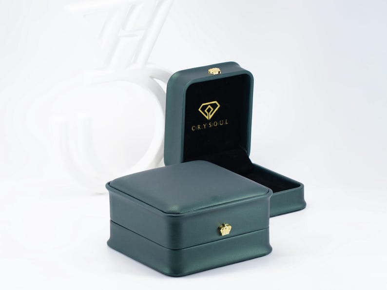 May include: A dark green square jewelry box with a smooth finish and a gold crown-shaped clasp on the front. The interior is lined with black velvet and features a gold diamond-shaped logo with the text CRYSOUL printed on the inside lid.