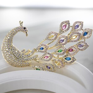 May include: A gold-coloured peacock brooch, adorned with sparkling clear and colourful gemstones. The peacock's body is covered in small, clear crystals, and its tail feathers feature various coloured gems. The brooch is a decorative accessory.