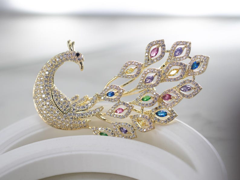 May include: A gold-coloured peacock brooch, adorned with sparkling clear and colourful gemstones. The peacock's body is covered in small, clear crystals, and its tail feathers feature various coloured gems. The brooch is a decorative accessory.