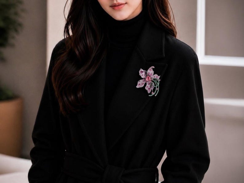 May include: A floral brooch featuring pink and purple petals with embedded crystals, accented by green curved stems. The brooch is pinned to the lapel of a black coat worn over a black turtleneck, creating a high-contrast accessory display.