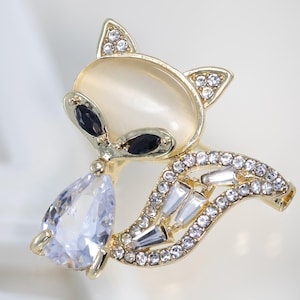 May include: A gold-toned fox brooch with a pearlescent body and black eyes. The fox's tail and ears are adorned with clear and white crystals. A large, teardrop-shaped, clear crystal hangs from the fox's chin.