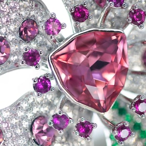 May include: A silver-toned brooch shaped like a flower, featuring a large central triangular pink gemstone surrounded by smaller round magenta stones and numerous tiny clear white crystals set into the metallic petals of the floral design.