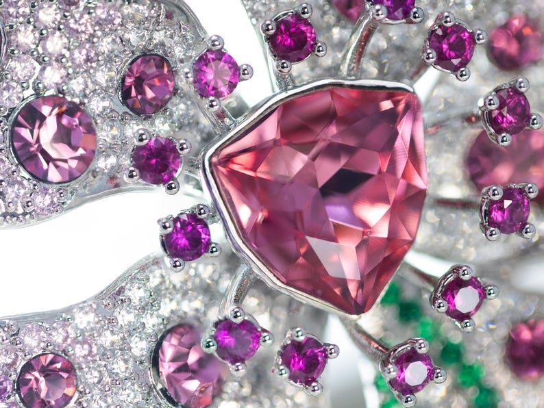 May include: A silver-toned brooch shaped like a flower, featuring a large central triangular pink gemstone surrounded by smaller round magenta stones and numerous tiny clear white crystals set into the metallic petals of the floral design.